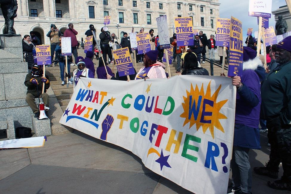Workers from various backgrounds gather to advocate for better work conditions, holding a banner that reads “What we could win together.”
